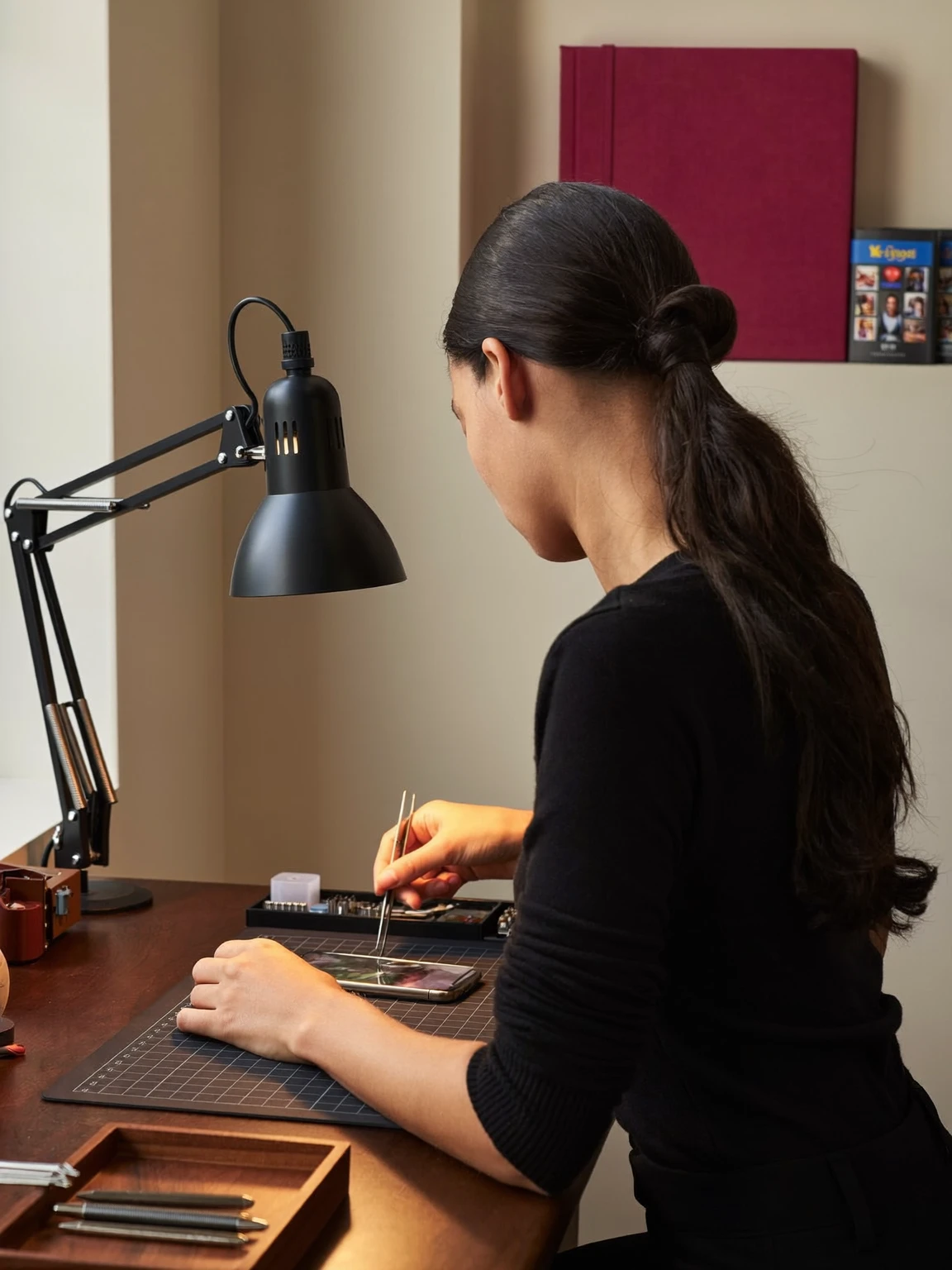 La fondatrice de Ravive Mémoire, de dos, à son établi : sous une lampe d’architecte, elle travaille avec une pince fine sur un téléphone posé sur un tapis de coupe.