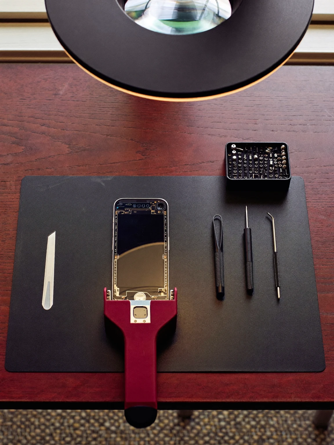 Établi de réparation vu de dessus : téléphone ouvert maintenu dans un support bourgogne, outils de précision et boîte de vis alignés sur un tapis noir, sous une lampe loupe.
