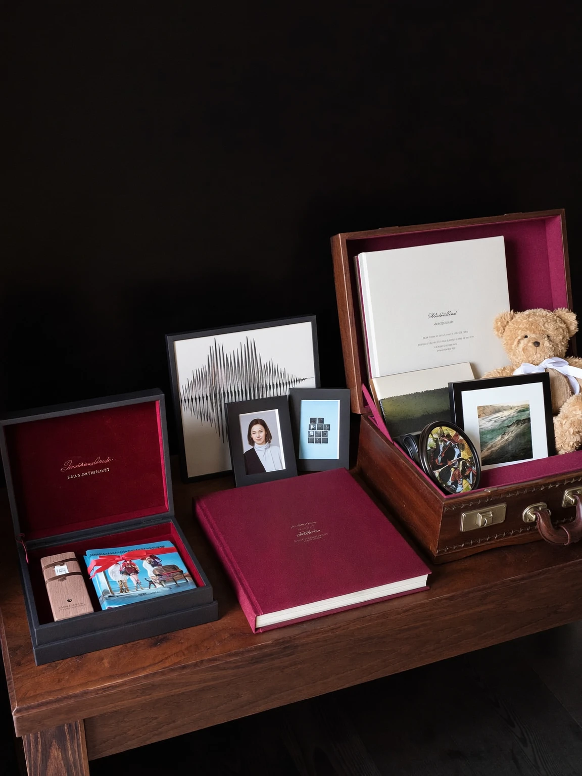 Les trois kits souvenirs sur une table en bois : un écrin noir avec clé USB et tirages, un album relié bourgogne, et un coffret en bois ouvert contenant un album, des cadres et un ourson.