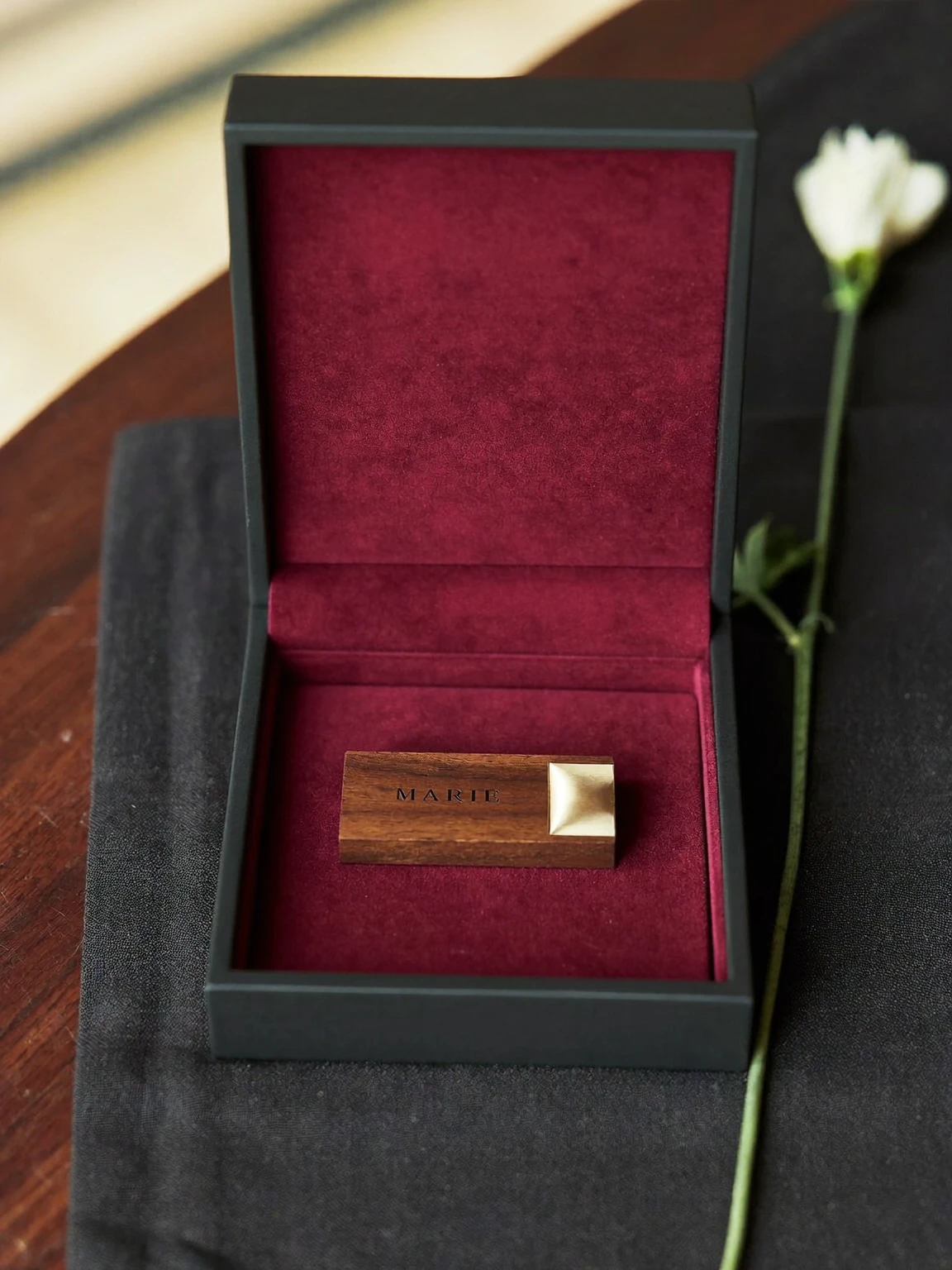 Écrin noir ouvert, doublé de velours bourgogne, contenant une clé USB en bois gravée « Marie », avec une fleur blanche posée à côté.