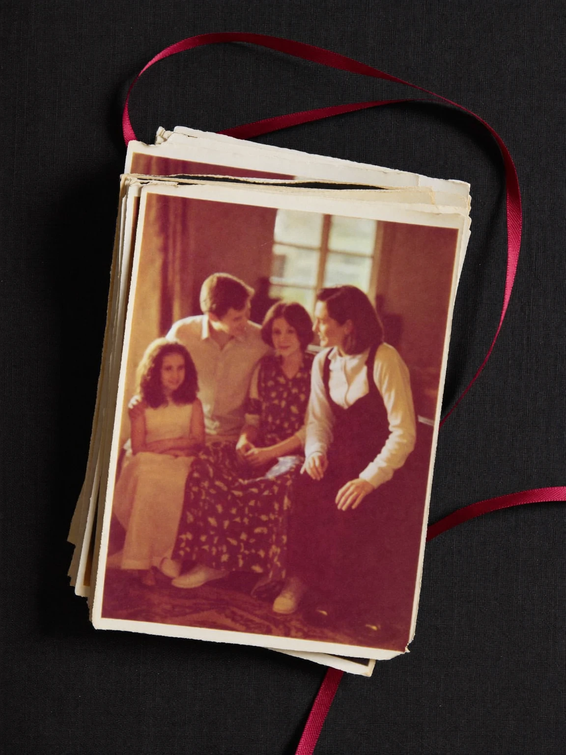 Pile de tirages photo aux tons chauds montrant une famille réunie, avec un ruban bourgogne, sur un tissu noir.