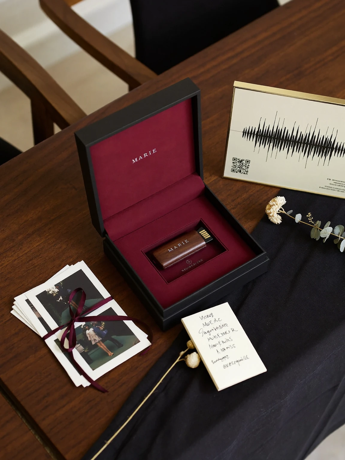 Écrin noir doublé de velours bourgogne contenant une clé USB en bois gravée « Marie », à côté de tirages photo noués d’un ruban et d’un cadre montrant l’onde d’une voix.