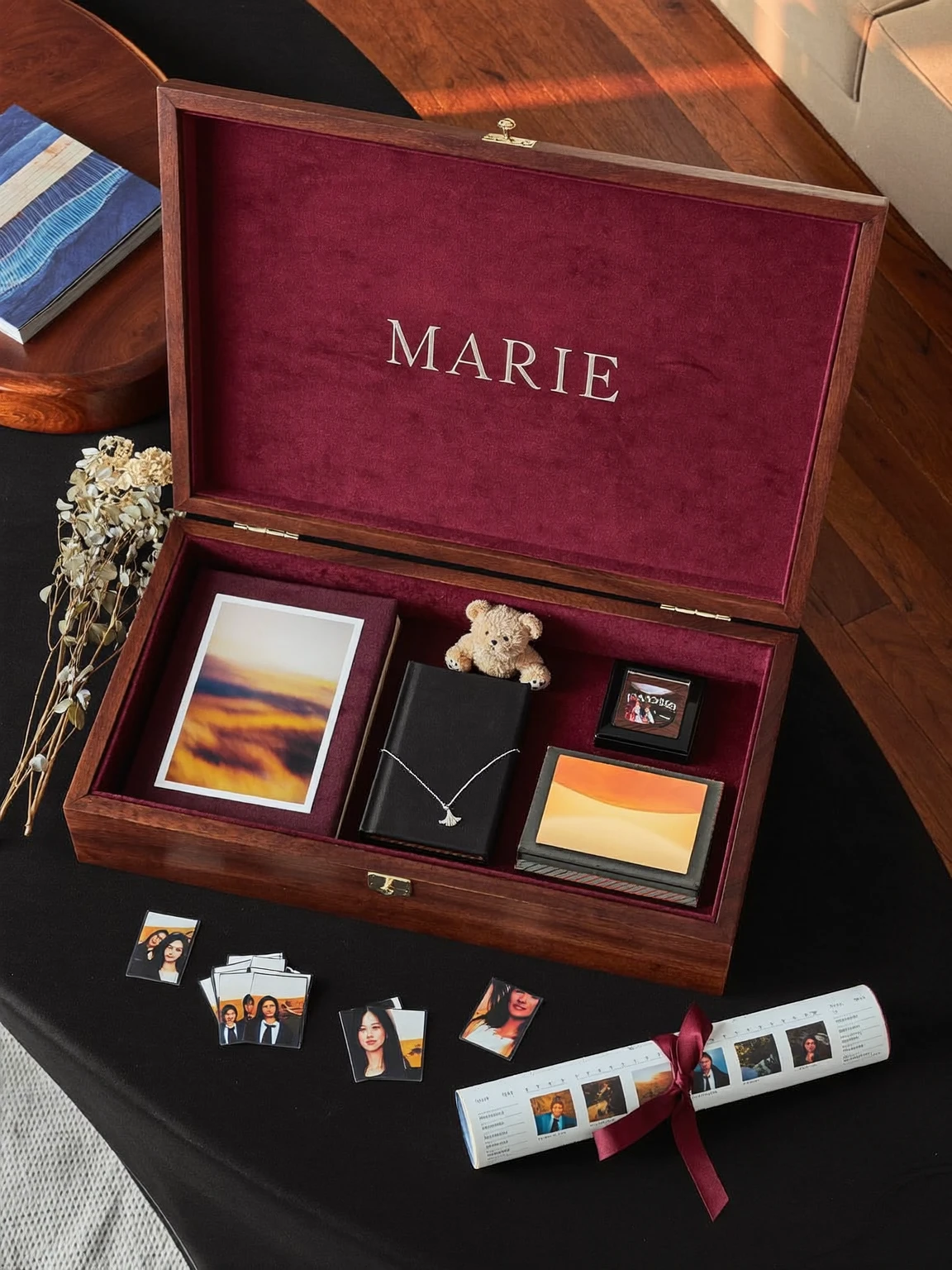 Coffret en bois ouvert, gravé « Marie » sur le velours bourgogne du couvercle, contenant tirages, livre, ourson et cadres, avec des photos et un rouleau noué d’un ruban à côté.